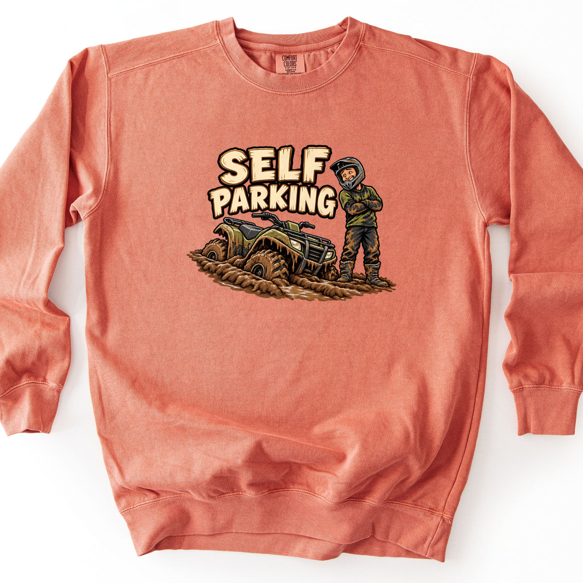 Self Parking Comfort Colors crewneck sweatshirt in Terracotta, laid flat. A cartoon rider in a helmet, green shirt and muddy pants stands with crossed arms beside a quad ATV stuck in mud, under the words Self Parking. Shown on a white background.