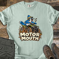 Motor Mouth Comfort Colors T-shirt in Bay, laid flat. A cartoon blue jay in a tan helmet squawks while riding a green four-wheeler through splattering mud above the words Motor Mouth. Folded olive pants and brown hiking boots sit on weathered wood planks.