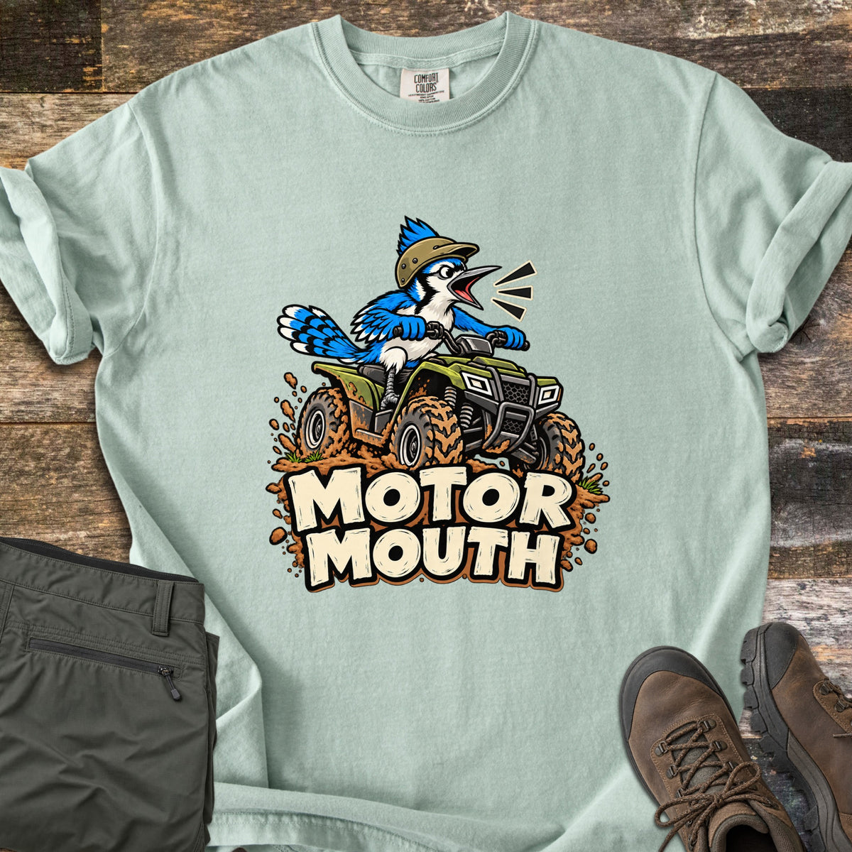 Motor Mouth Comfort Colors T-shirt in Bay, laid flat. A cartoon blue jay in a tan helmet squawks while riding a green four-wheeler through splattering mud above the words Motor Mouth. Folded olive pants and brown hiking boots sit on weathered wood planks.