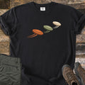 Flying Clays Comfort Colors T-shirt in Black, laid flat. Three clay pigeon targets in orange, olive green and cream fly diagonally upward across the chest with motion trails. Folded green cargo pants and brown hiking boots sit beside it on weathered wood planks.