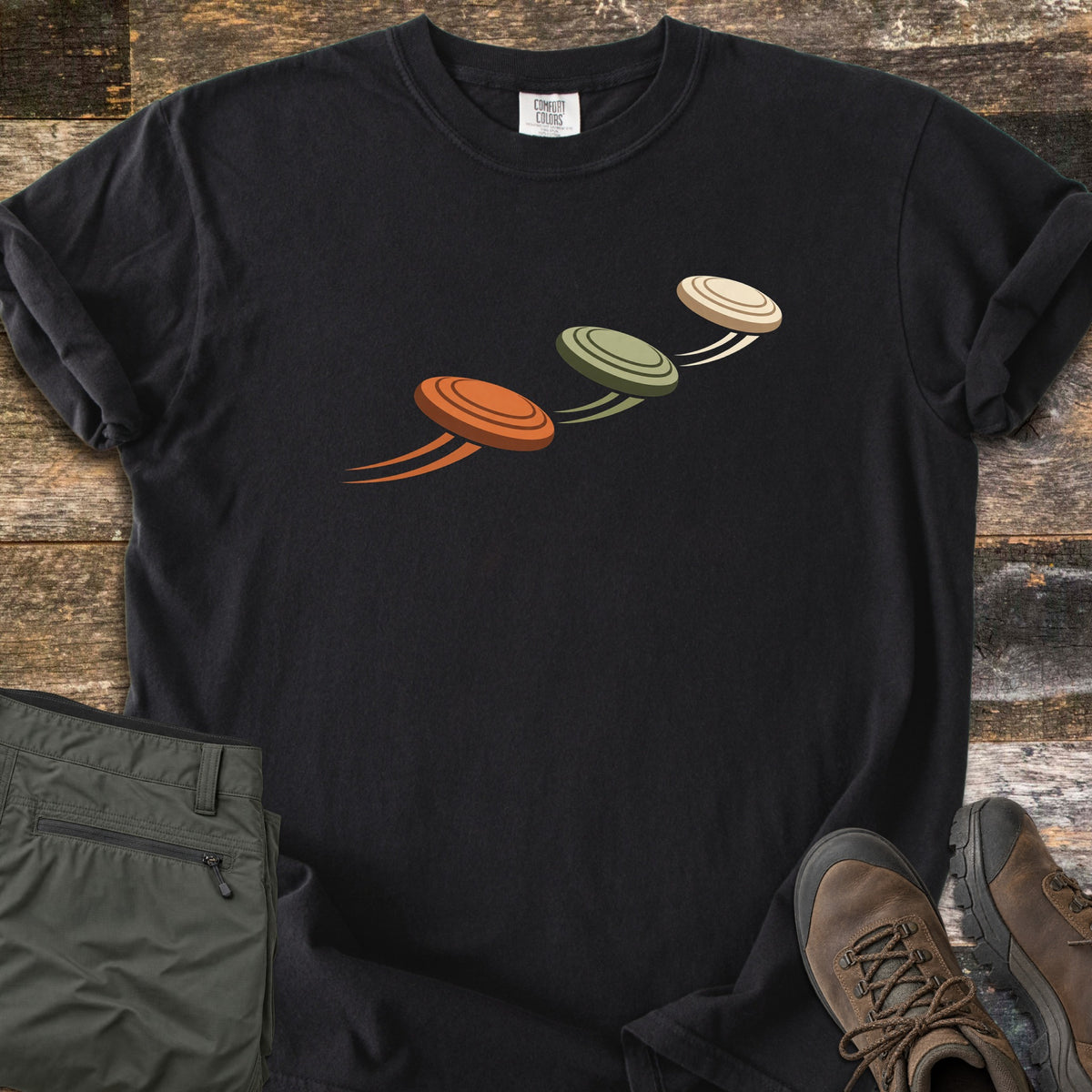 Flying Clays Comfort Colors T-shirt in Black, laid flat. Three clay pigeon targets in orange, olive green and cream fly diagonally upward across the chest with motion trails. Folded green cargo pants and brown hiking boots sit beside it on weathered wood planks.