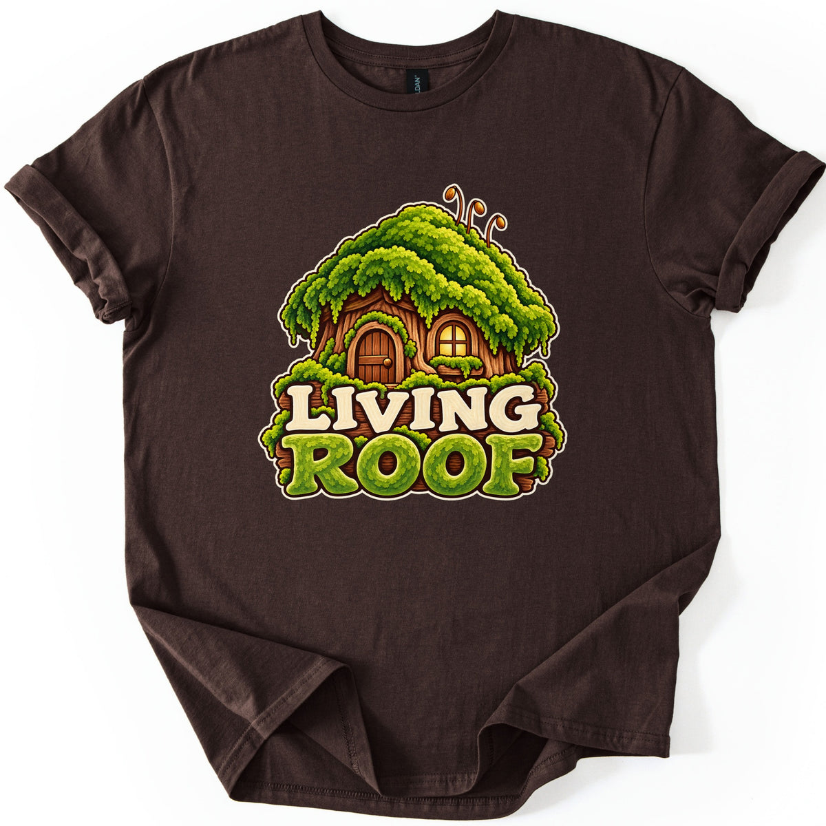 Living Roof Comfort Colors T-shirt in Dark Chocolate, laid flat. A cartoon tree-stump cottage with a thick green moss roof, an arched wooden door and a glowing window sits above the words Living Roof in cream and moss-green letters. Sleeves are cuffed on a white surface.