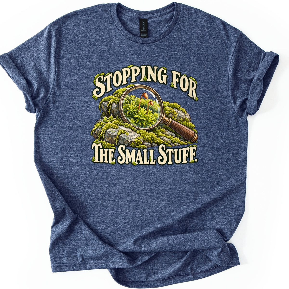 Stopping for the Small Stuff Comfort Colors T-shirt in Heather Navy, laid flat. A wooden-handled magnifying glass rests on moss-covered rocks, its lens enlarging moss with tiny spore capsules, between the words Stopping For The Small Stuff. Sleeves are rolled on a white background.