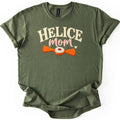 Helice Mom Comfort Colors T-shirt in Military Green, laid flat. Cream arched letters read HELICE above pink script reading mom with a small heart, over an orange two-bladed helice target with a cream center ring. The shirt has rolled sleeves on a white background.