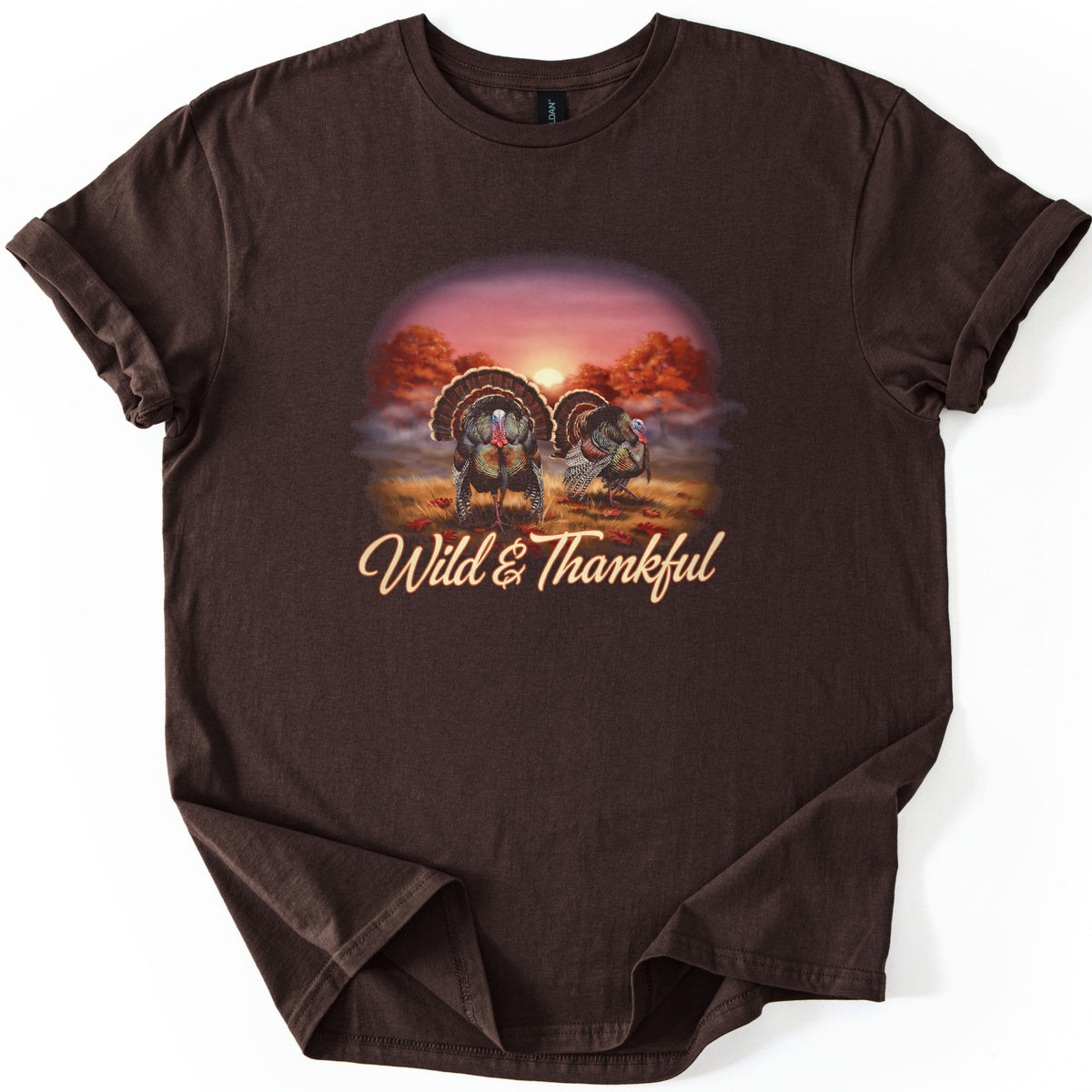 Wild & Thankful Comfort Colors T-shirt in Dark Chocolate, laid flat. Two wild turkeys with fanned tails stand in a grassy field under orange autumn trees at sunset, above the script words Wild & Thankful. The sleeves are rolled, on a white background.