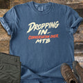 Dropping In Conversation Over Comfort Colors T-shirt in Denim, laid flat. Cream brush-stroke letters read Dropping In- and MTB, with Conversation Over in orange-red between them. Sleeves are cuffed on weathered wood planks beside folded olive pants and brown hiking boots.