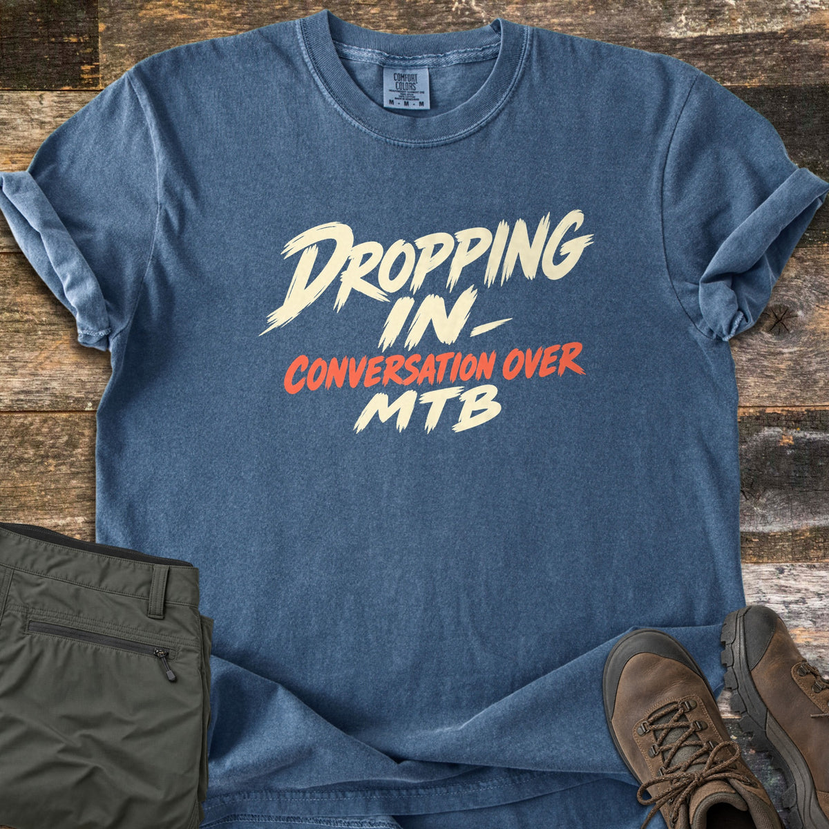 Dropping In Conversation Over Comfort Colors T-shirt in Denim, laid flat. Cream brush-stroke letters read Dropping In- and MTB, with Conversation Over in orange-red between them. Sleeves are cuffed on weathered wood planks beside folded olive pants and brown hiking boots.