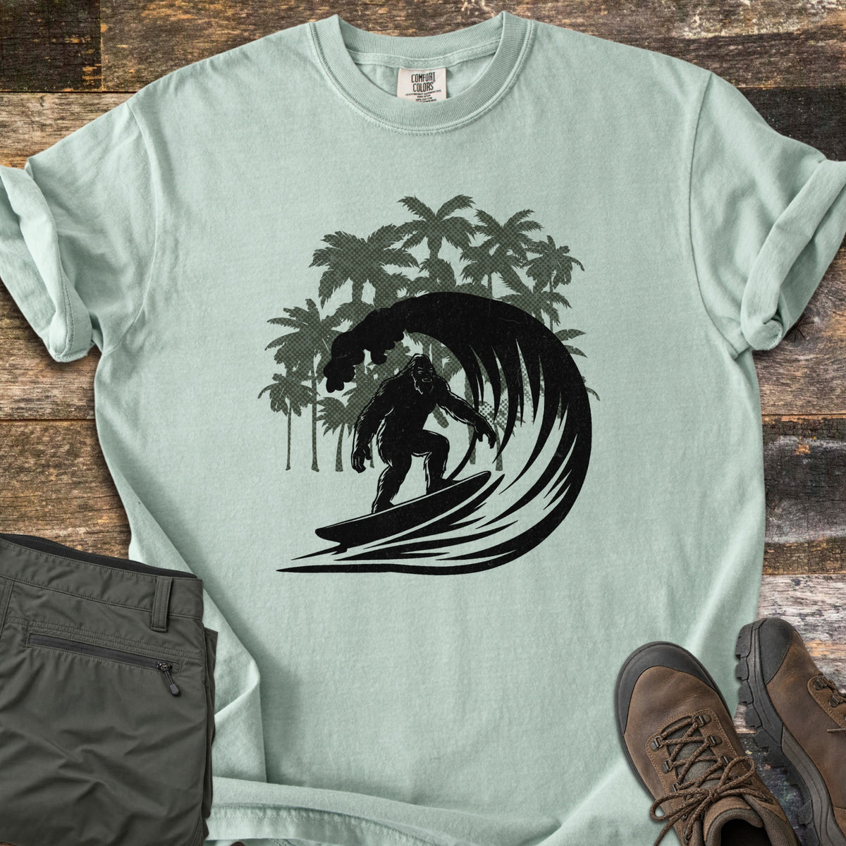 Surf Squatch Comfort Colors T-shirt in Bay, laid flat. A black silhouette of Bigfoot surfing inside a curling wave, with green palm trees behind, and no words. The shirt lies on weathered wood with folded olive pants and brown hiking boots.