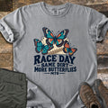 Race Day Same Dirt More Butterflies Comfort Colors T-shirt in Grey, laid flat. Three teal and orange butterflies over a cream race number plate with a tire track, above the words Race Day Same Dirt More Butterflies MTB. Folded olive pants and brown hiking boots sit on weathered wood.