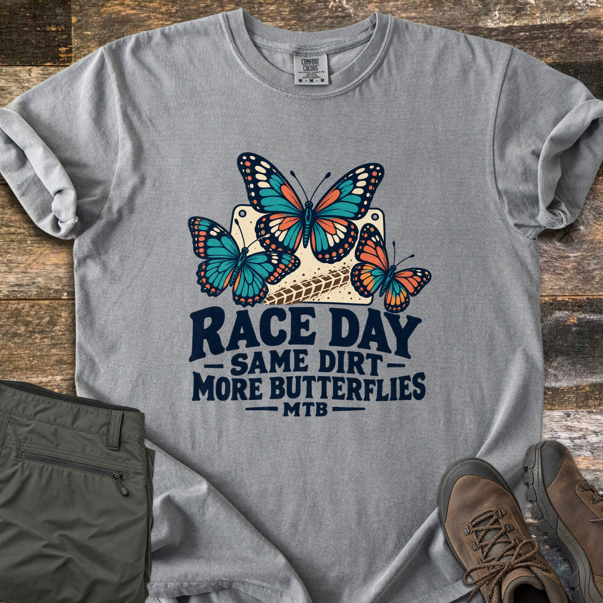 Race Day Same Dirt More Butterflies Comfort Colors T-shirt in Grey, laid flat. Three teal and orange butterflies over a cream race number plate with a tire track, above the words Race Day Same Dirt More Butterflies MTB. Folded olive pants and brown hiking boots sit on weathered wood.