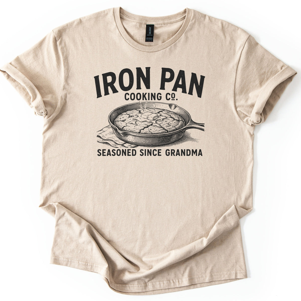 Iron Pan Cooking Co Comfort Colors T-shirt in Sand, laid flat. Black print of the words Iron Pan Cooking Co. above an engraved-style cast iron skillet of cornbread topped with melting butter on a cloth, with Seasoned Since Grandma below. Cuffed sleeves on a white background.