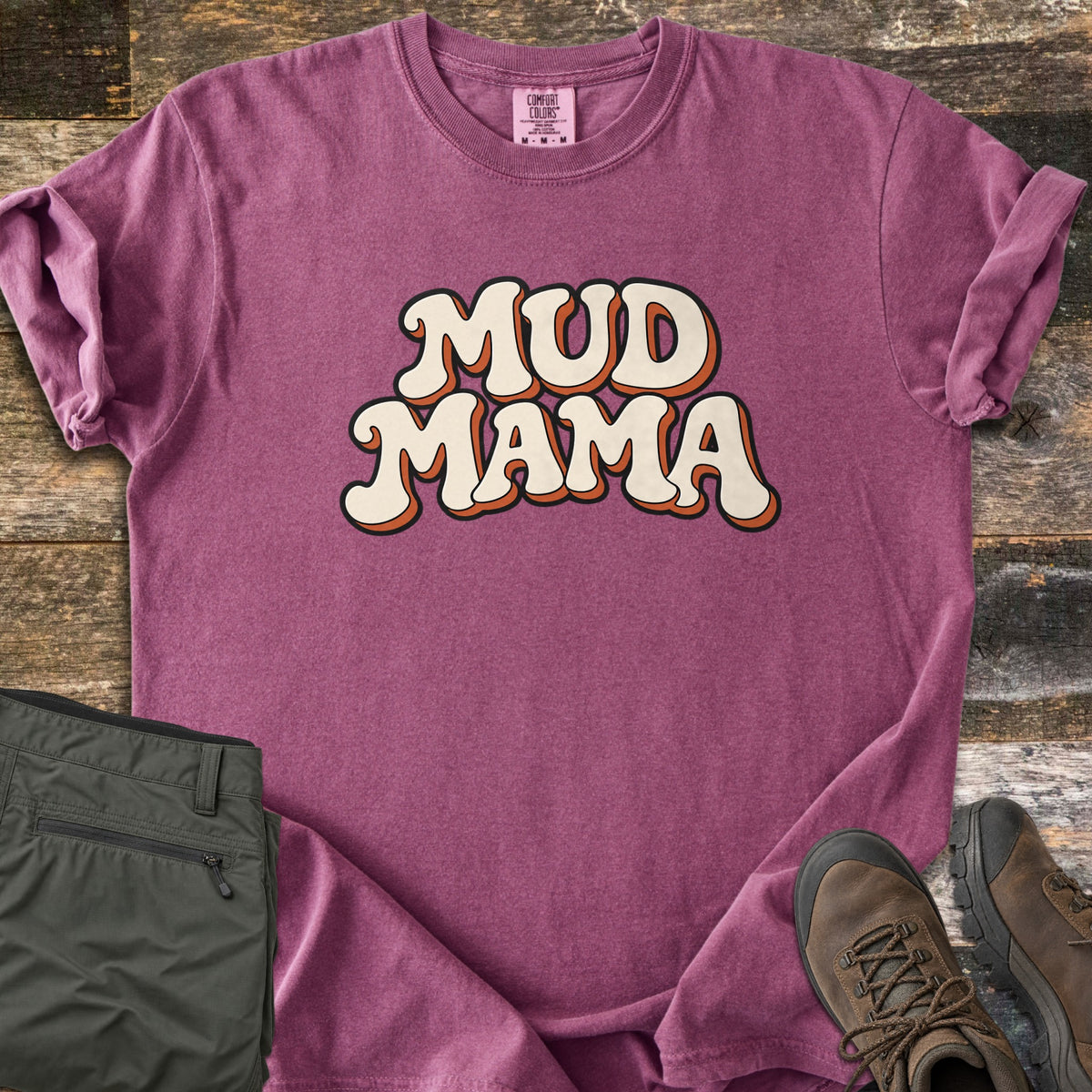 Mud Mama Comfort Colors T-shirt in Berry, laid flat. The words MUD MAMA are printed in cream retro bubble letters with an orange drop shadow and black outline. The shirt lies on weathered wood planks beside folded olive hiking pants and brown hiking boots.