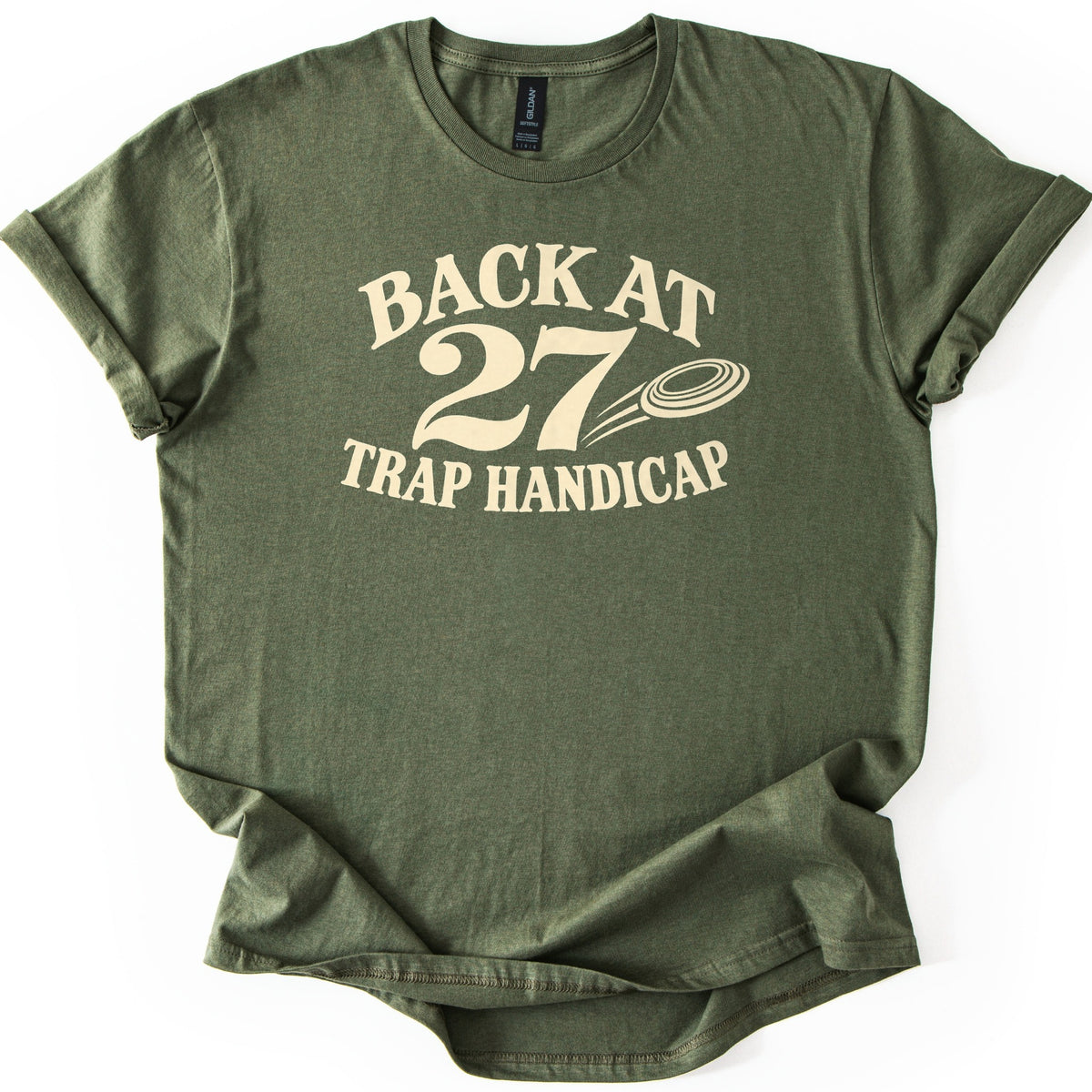 Back at 27 Trap Handicap Comfort Colors T-shirt in Military Green, laid flat. Cream lettering reads Back at 27 Trap Handicap, with a clay target trailing motion lines beside the large 27. The shirt has rolled sleeves and lies on a white background.