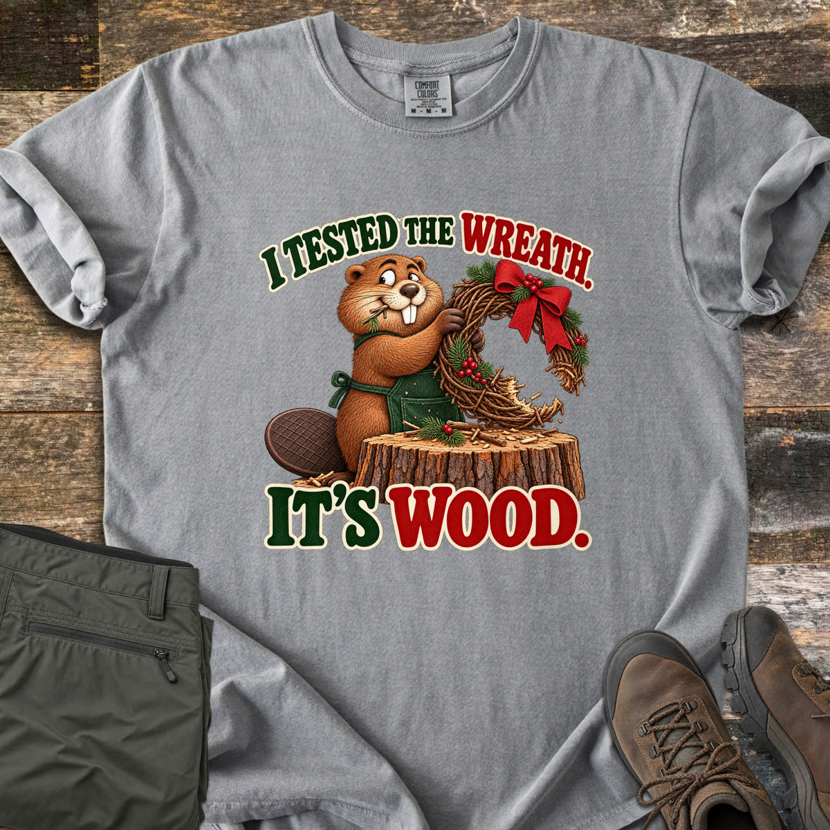 I Tested the Wreath. It's Wood. Comfort Colors T-shirt in Grey, laid flat. A cartoon beaver in a green apron chews a twig wreath with a red bow on a tree stump, under the words I Tested the Wreath. It's Wood. Olive pants and brown hiking boots lie beside it on weathered wood planks.