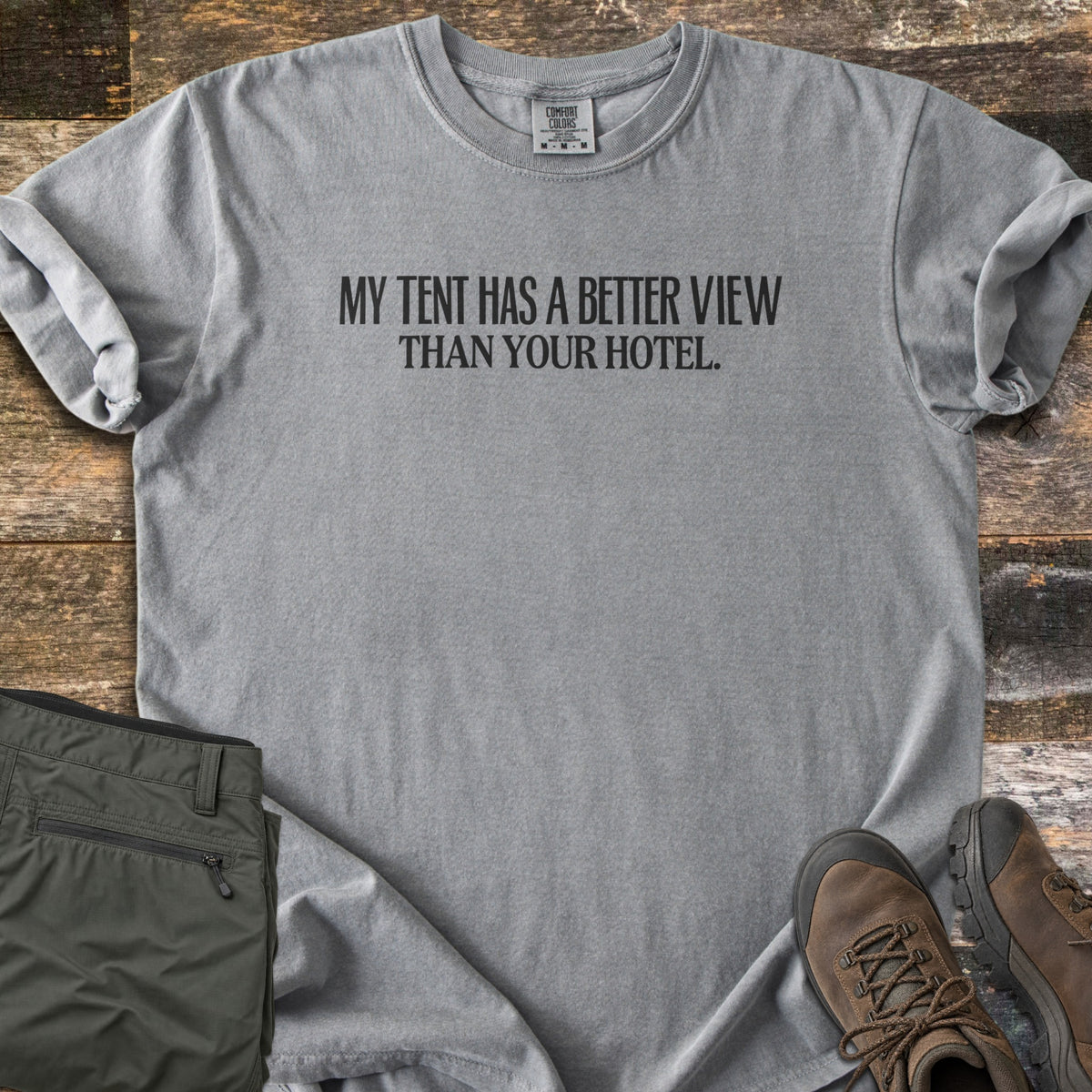 Better View Than Your Hotel Comfort Colors T-shirt in Grey, laid flat. Black text reads My Tent Has a Better View Than Your Hotel. Sleeves are cuffed, on a weathered wood surface with folded olive hiking pants and brown hiking boots.