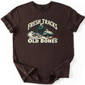 Fresh Tracks Old Bones Comfort Colors T-shirt in Dark Chocolate, laid flat. A skeleton in a white helmet and teal jacket rides a vintage teal, white and orange snowmobile through spraying snow, between the words Fresh Tracks and Old Bones. Sleeves are cuffed on a white background.