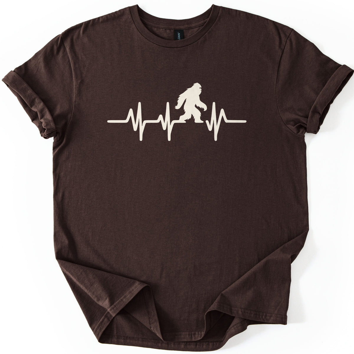 Yeti Heartbeat Comfort Colors T-shirt in Dark Chocolate, laid flat. A cream-colored heartbeat line runs across the chest, with a walking Bigfoot silhouette standing within it; there are no words. The sleeves are cuffed and the shirt lies on a white background.