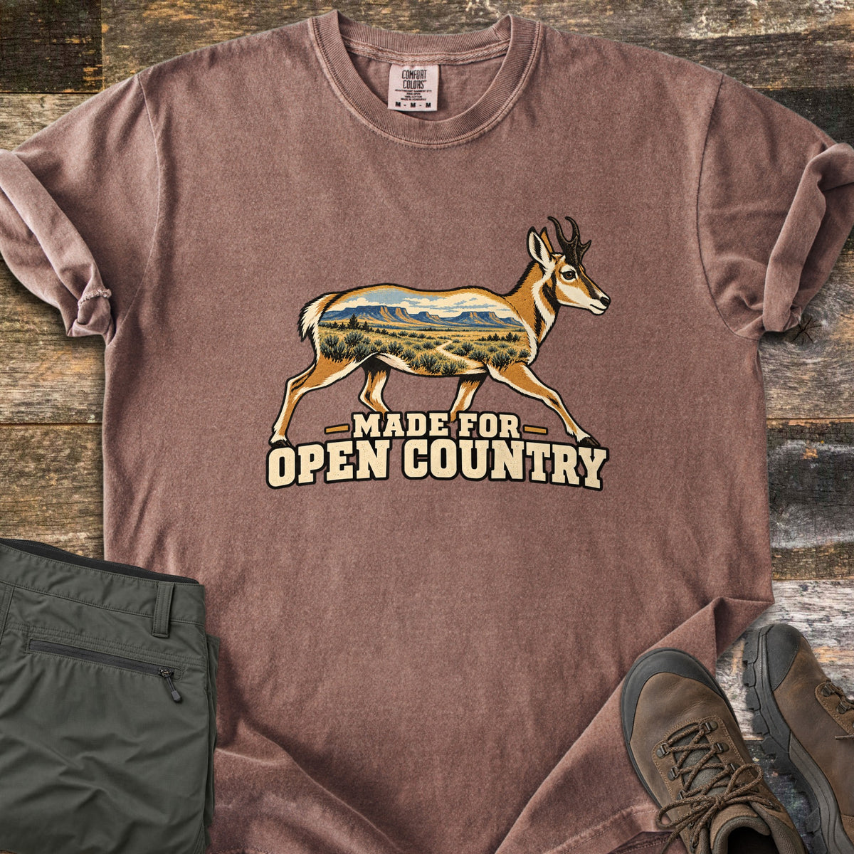 Made for Open Country Comfort Colors T-shirt in Espresso, laid flat. A running pronghorn whose body is filled with a desert scene of mesas, sagebrush and a trail, above the words Made for Open Country. Sleeves are rolled on a wood background with green pants and brown hiking boots.