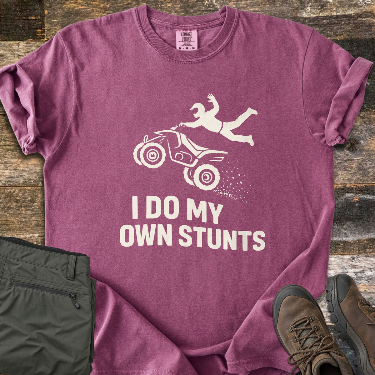 ATV Stunts Comfort Colors T-shirt in Berry, laid flat. A white silhouette of a helmeted rider flying off a wheelie-popping ATV kicking up dirt, above the words I Do My Own Stunts. Olive cargo pants and brown hiking boots sit beside it on weathered wood.