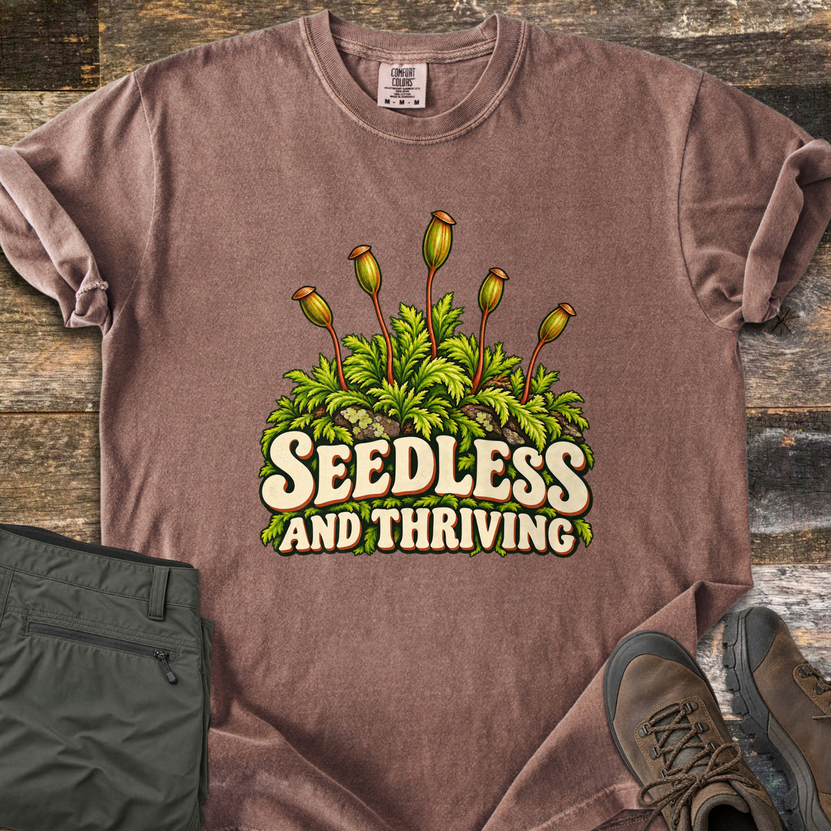 Seedless and Thriving Comfort Colors T-shirt in Espresso, laid flat. A mound of green moss with tall reddish stalks topped by spore capsules sits above the words Seedless and Thriving in cream retro letters. It lies on weathered wood with folded olive pants and brown hiking boots.