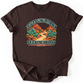 Desert Singletrack Prickly Company Comfort Colors T-shirt in Dark Chocolate, laid flat. Arched words Desert Singletrack top a dirt trail winding between prickly pear cacti and red rock buttes, with Prickly Company and MTB below. The shirt lies on a white surface with cuffed sleeves.
