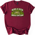 Hide & Seek Moss Edition Comfort Colors T-shirt in Maroon, laid flat. A wide-eyed spotted green frog peeks out of a bed of bright green moss between the cream words Hide & Seek and Moss Edition. The shirt has rolled sleeves and lies on a white background.