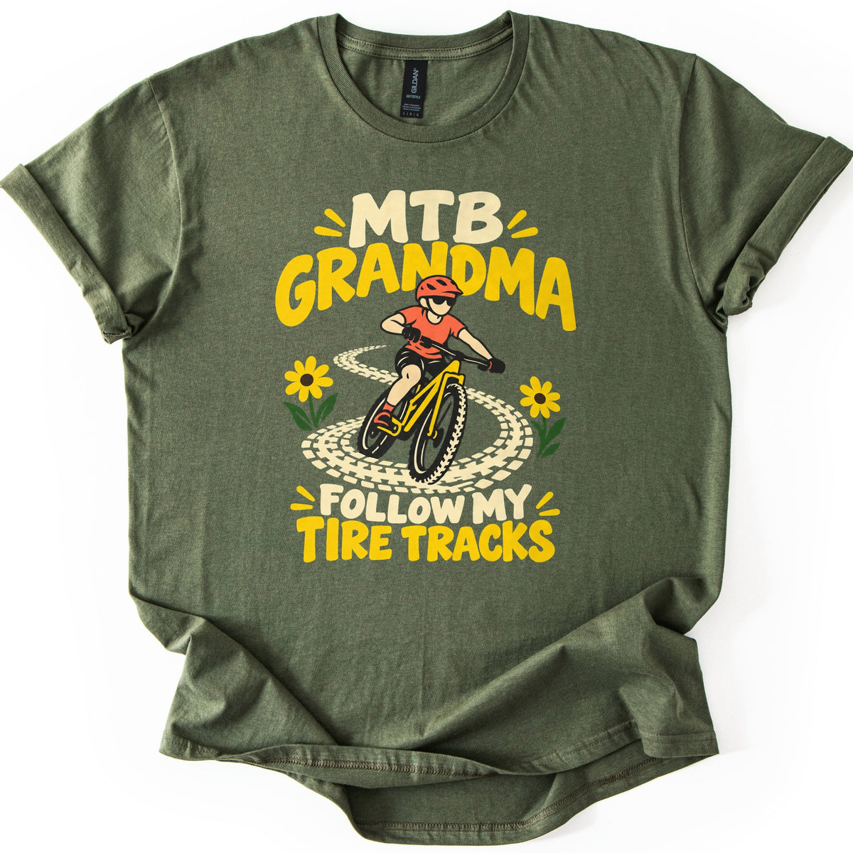 MTB Grandma Follow My Tire Tracks Comfort Colors T-shirt in Military Green, laid flat. A child in a red helmet and sunglasses rides a yellow mountain bike along looping tire tracks between yellow flowers, under MTB Grandma and above Follow My Tire Tracks. Sleeves rolled, on a white background.