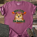 Wide Eyed for Doubles Comfort Colors T-shirt in Berry, laid flat. A wide-eyed cartoon owl sits between two orange clay targets under the arched words Wide Eyed, with For Doubles and Skeet below. Sleeves are rolled, on weathered wood with olive pants and brown hiking boots.