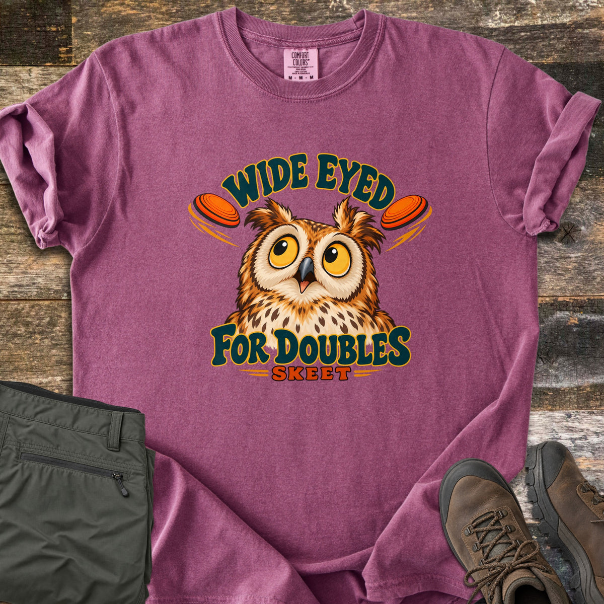 Wide Eyed for Doubles Comfort Colors T-shirt in Berry, laid flat. A wide-eyed cartoon owl sits between two orange clay targets under the arched words Wide Eyed, with For Doubles and Skeet below. Sleeves are rolled, on weathered wood with olive pants and brown hiking boots.