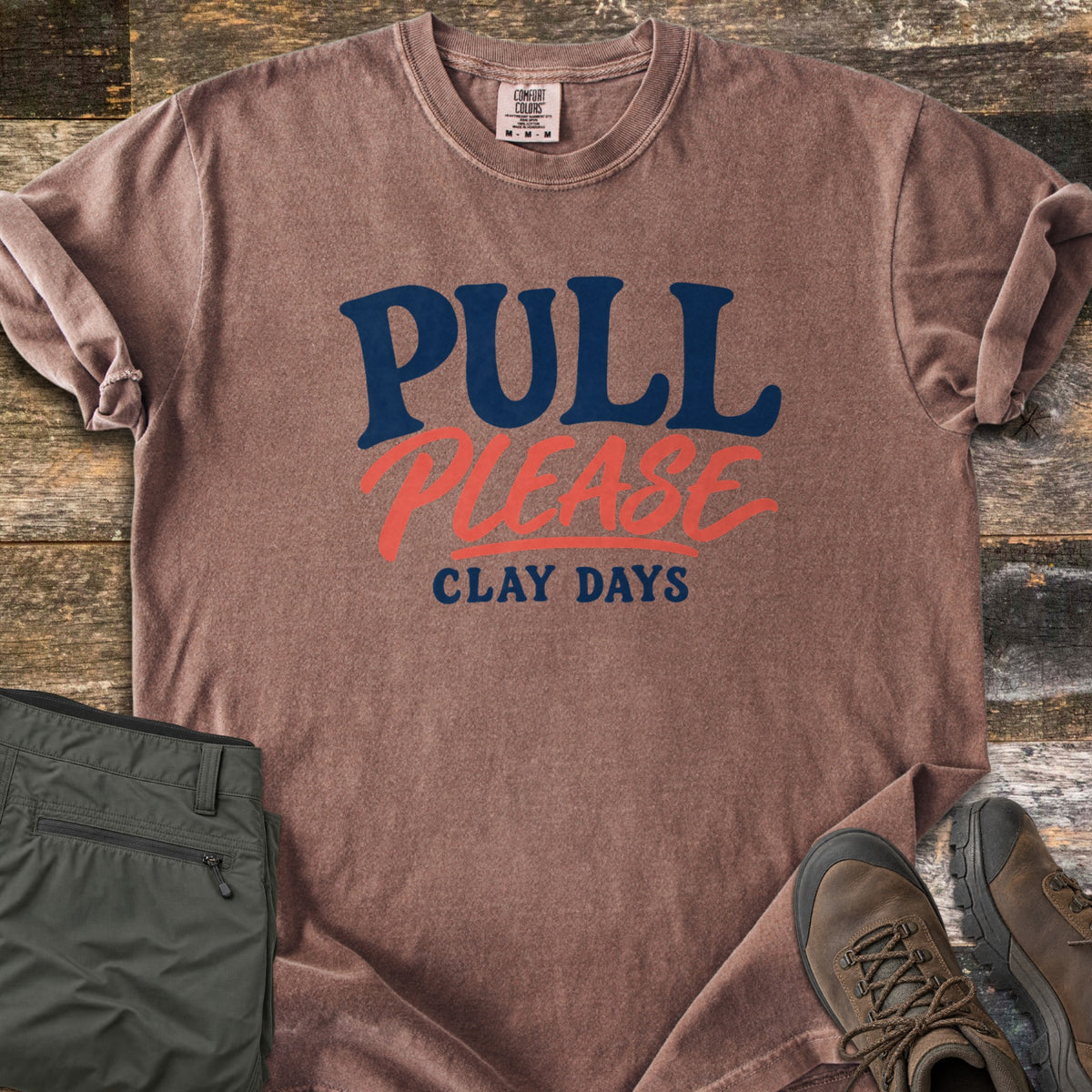 Pull Please Comfort Colors T-shirt in Espresso, laid flat. The words Pull Please Clay Days are printed in navy and coral lettering, with Please in coral script. The shirt lies on weathered wood with rolled sleeves, beside folded olive pants and brown hiking boots.