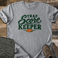 Trap Score Keeper Comfort Colors T-shirt in Grey, laid flat. Dark green lettering reads Trap Score Keeper above an orange clay target. Sleeves are cuffed on weathered wood, with folded olive cargo pants and brown hiking boots beside it.