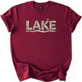 Lake Comfort Colors T-shirt in Maroon, laid flat. The word LAKE is spelled in weathered driftwood planks, with a small blue and orange kingfisher perched on the K and wavy water lines below. The shirt lies on white with its sleeves rolled.