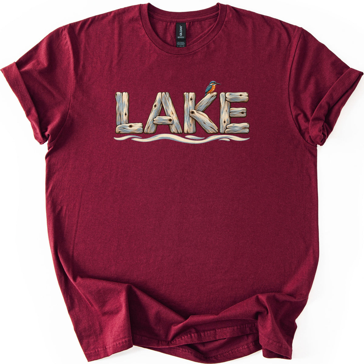 Lake Comfort Colors T-shirt in Maroon, laid flat. The word LAKE is spelled in weathered driftwood planks, with a small blue and orange kingfisher perched on the K and wavy water lines below. The shirt lies on white with its sleeves rolled.