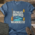 Human Powered Wanderlust Comfort Colors T-shirt in Denim, laid flat. A wooden canoe paddle and a yellow kayak paddle frame a winding turquoise river and the words Human Powered Wanderlust in cream. Folded olive pants and brown hiking boots sit beside it on weathered wood planks.