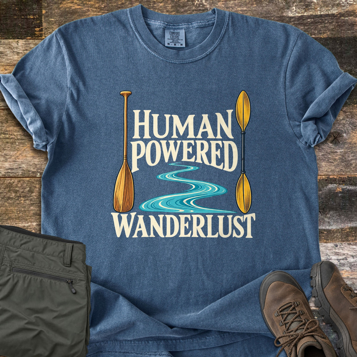 Human Powered Wanderlust Comfort Colors T-shirt in Denim, laid flat. A wooden canoe paddle and a yellow kayak paddle frame a winding turquoise river and the words Human Powered Wanderlust in cream. Folded olive pants and brown hiking boots sit beside it on weathered wood planks.