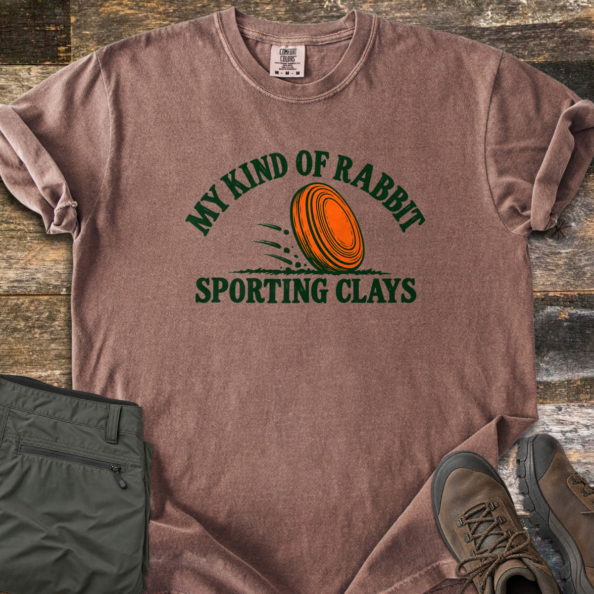 My Kind of Rabbit Comfort Colors T-shirt in Espresso, laid flat. An orange clay target rolls through grass between the curved words My Kind of Rabbit and Sporting Clays in dark green. Olive hiking pants and brown boots lie beside it on weathered wood planks.