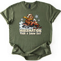 Hibernation Took a Snow Day Comfort Colors T-shirt in Military Green, laid flat. A grinning cartoon bear in a helmet and yellow scarf rides a red snowmobile through spraying snow under snowflakes, above the words Hibernation Took a Snow Day. The shirt lies on a white surface.
