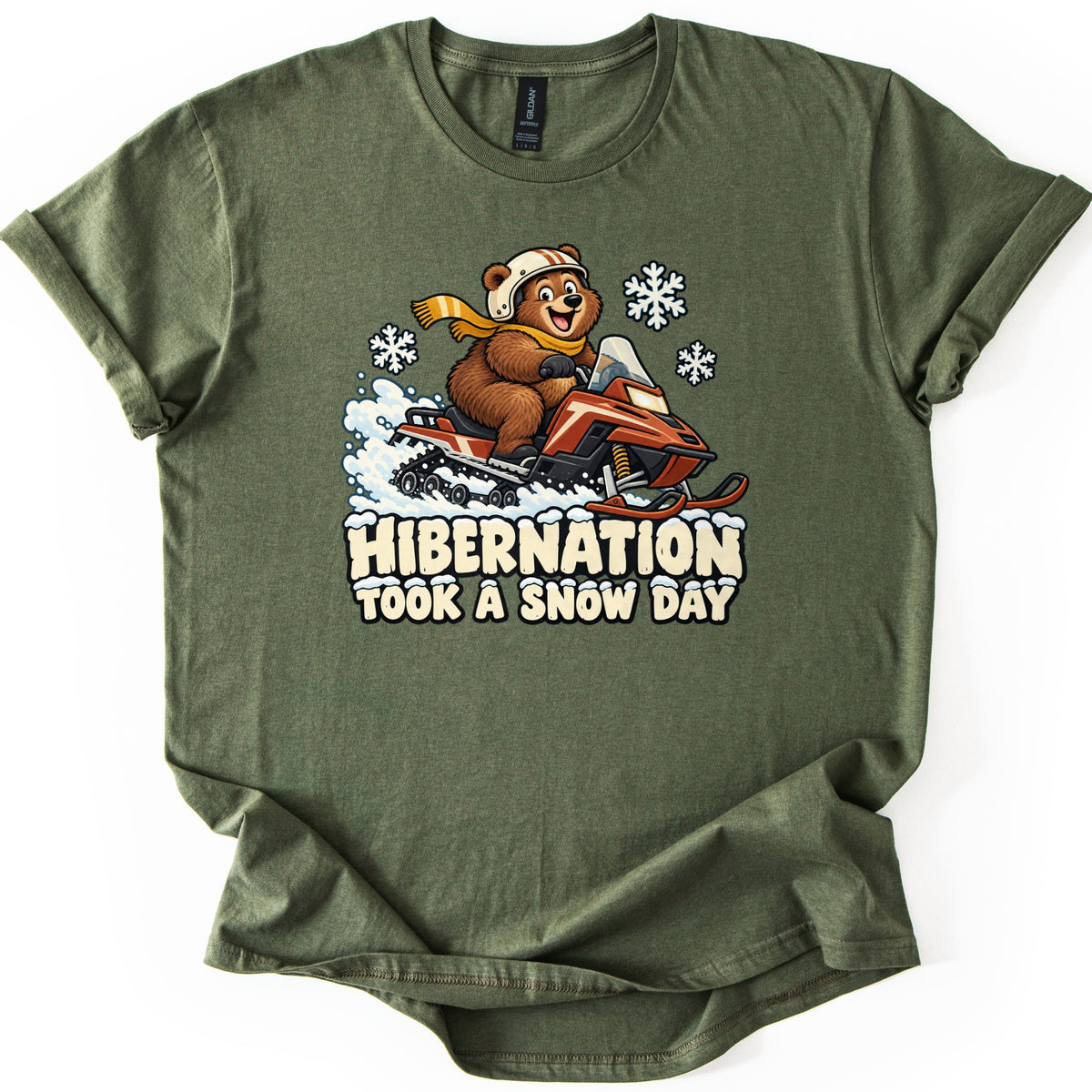 Hibernation Took a Snow Day Comfort Colors T-shirt in Military Green, laid flat. A grinning cartoon bear in a helmet and yellow scarf rides a red snowmobile through spraying snow under snowflakes, above the words Hibernation Took a Snow Day. The shirt lies on a white surface.