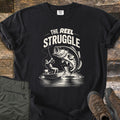 Reel Struggle Comfort Colors T-shirt in Black, laid flat. A white print of a bass leaping from the water toward a man reeling it in from a small boat, under the words The Reel Struggle. Olive cargo pants and brown hiking boots lie beside it on dark wood planks.