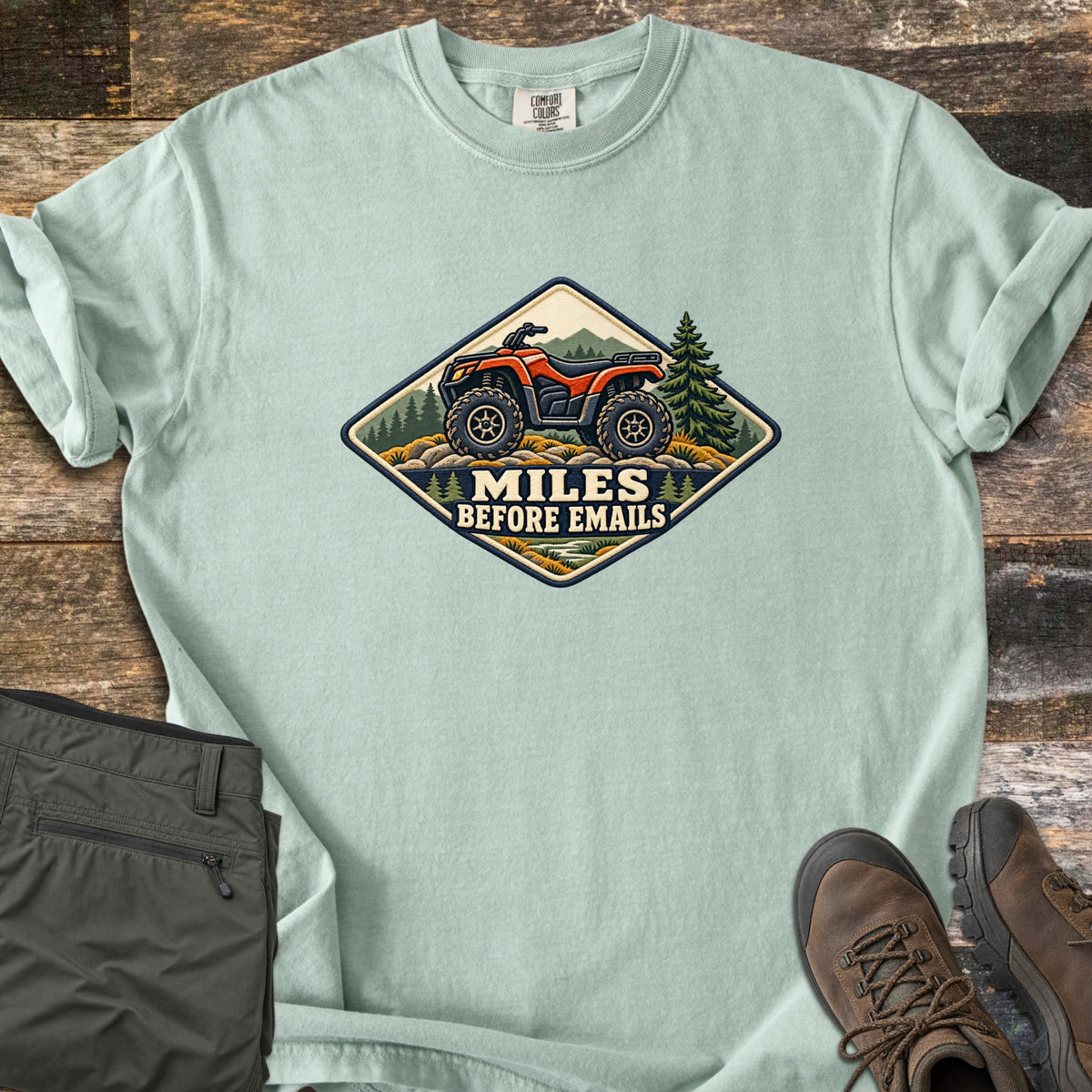 Miles Before Emails Comfort Colors T-shirt in Bay, laid flat. A diamond-shaped patch shows a red ATV on rocks with pine trees and mountains above the words Miles Before Emails. Sleeves rolled, on weathered wood with green pants and brown hiking boots.