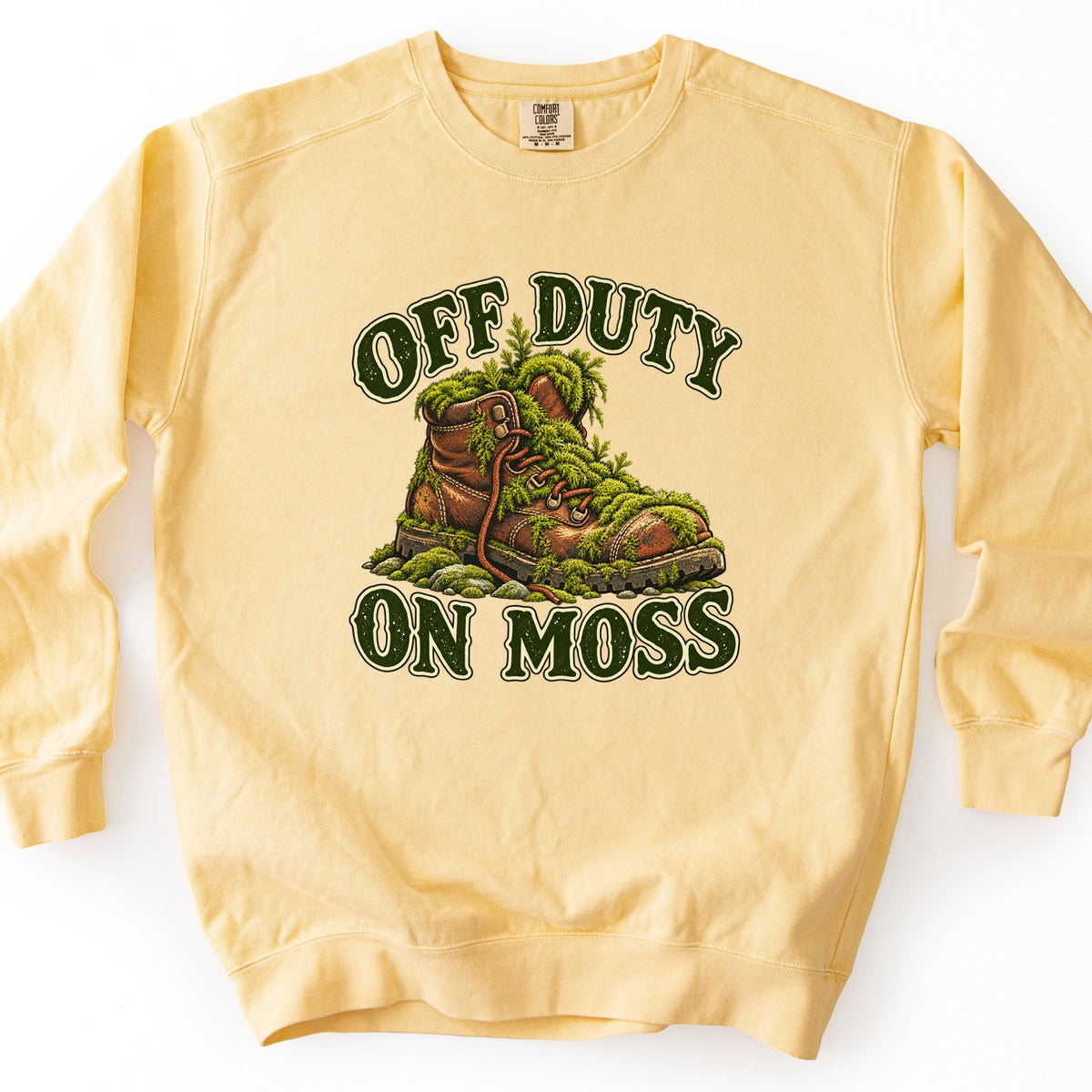 Off Duty on Moss Comfort Colors crewneck sweatshirt in Butter, laid flat. A brown leather hiking boot overgrown with green moss and ferns sits between the words Off Duty and On Moss in dark green arched lettering. The sweatshirt lies on a white surface.