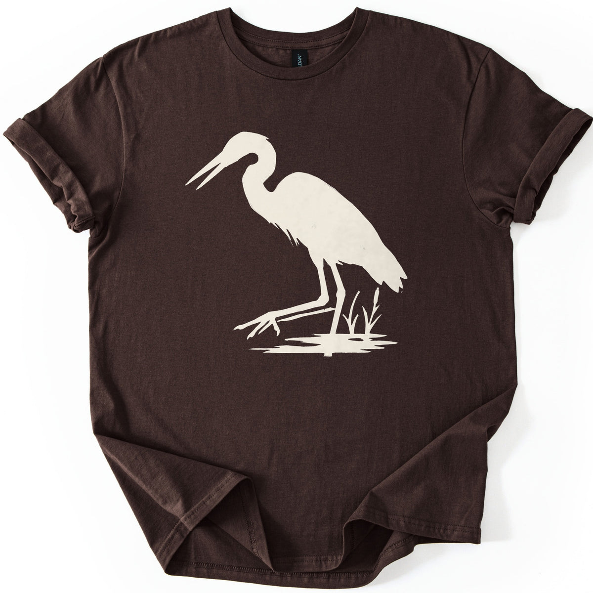 Great Blue Heron Comfort Colors T-shirt in Dark Chocolate, laid flat. A cream silhouette of a heron wading in shallow water beside cattails, beak open and one foot reaching forward. The shirt lies on a white surface with cuffed sleeves.
