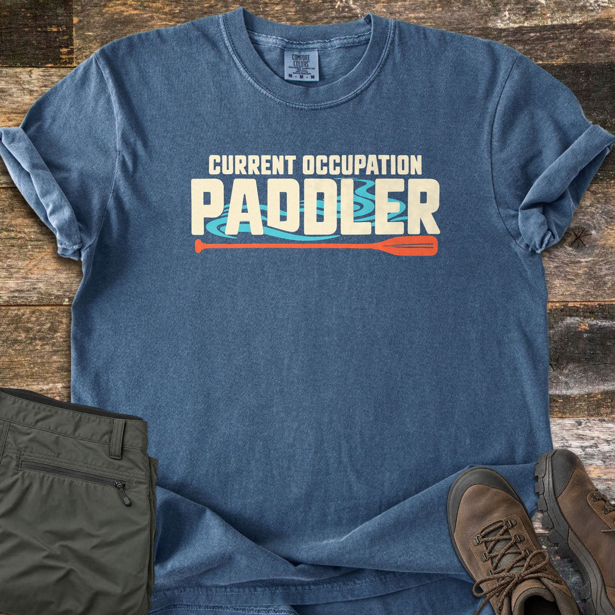 Current Occupation Paddler Comfort Colors T-shirt in Denim, laid flat. Cream block letters read Current Occupation Paddler over turquoise water swirls, with an orange canoe paddle beneath. Sleeves are rolled on a weathered wood surface beside olive hiking pants and brown hiking boots.