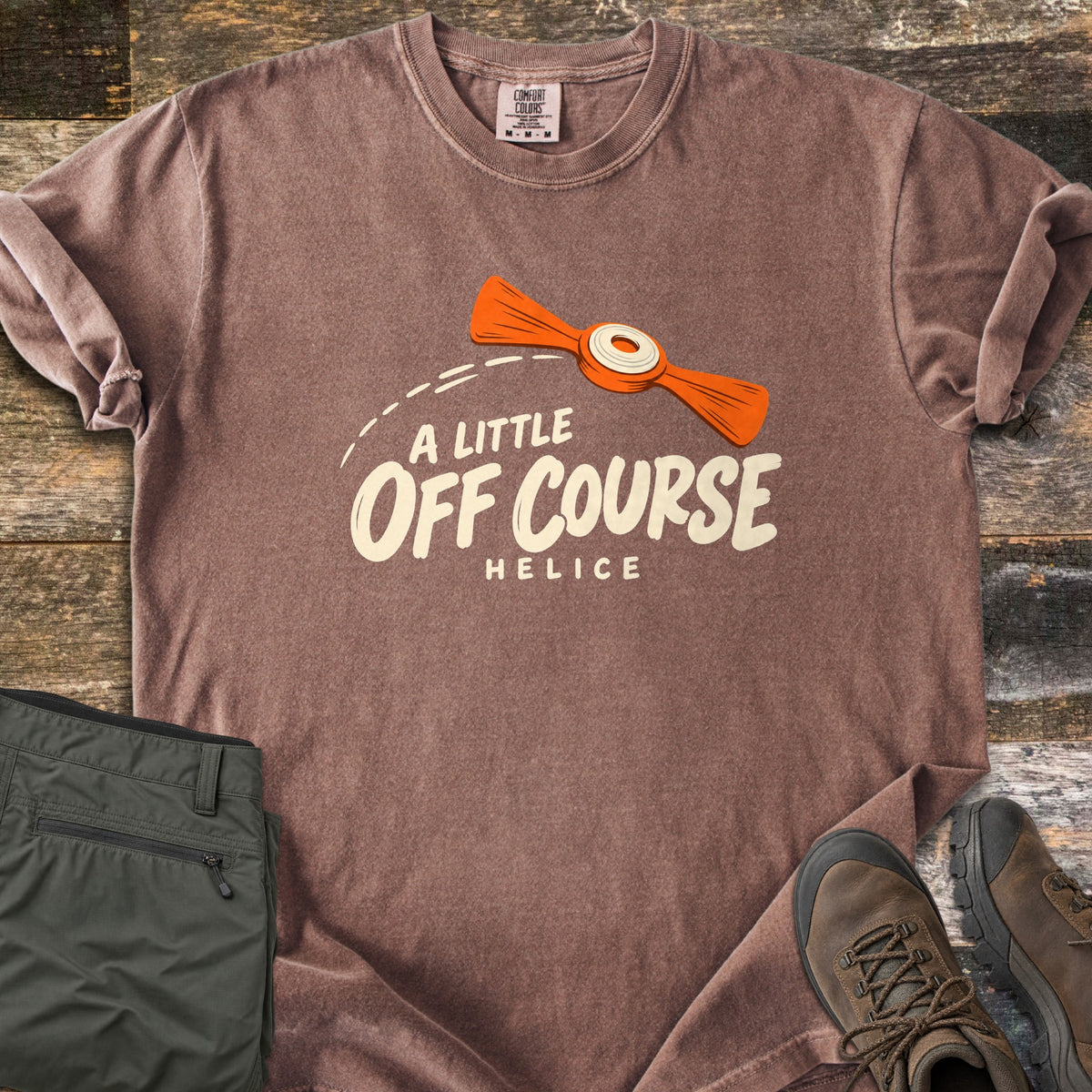 A Little Off Course Comfort Colors T-shirt in Espresso, laid flat. An orange helice target with a white center flies along a dashed arc above cream text reading A Little Off Course and Helice. Folded olive hiking pants and brown hiking boots sit beside it on weathered wood.