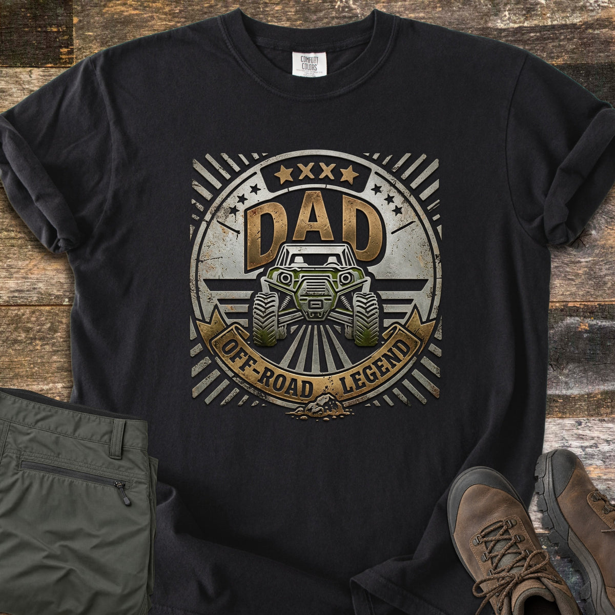 Dad Off Road Legend Comfort Colors T-shirt in Black, laid flat. A distressed metallic badge shows a green off-road UTV under the word DAD, with stars and a banner reading Off-Road Legend. Olive cargo pants and brown hiking boots sit beside it on weathered wood.