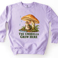 The Umbrella Grew Here Comfort Colors crewneck sweatshirt in Orchid, laid flat. A mouse wearing a green backpack shelters under a large orange mushroom as raindrops fall, beside a puddle, ferns, and moss, above the words The Umbrella Grew Here in dark green. Shown on a white background.