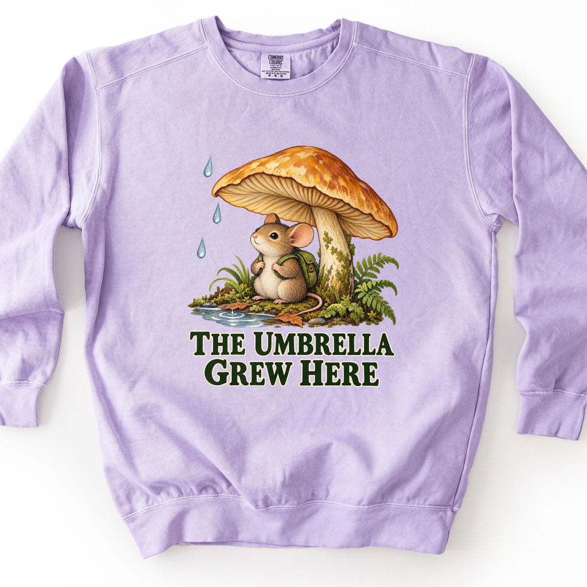The Umbrella Grew Here Comfort Colors crewneck sweatshirt in Orchid, laid flat. A mouse wearing a green backpack shelters under a large orange mushroom as raindrops fall, beside a puddle, ferns, and moss, above the words The Umbrella Grew Here in dark green. Shown on a white background.