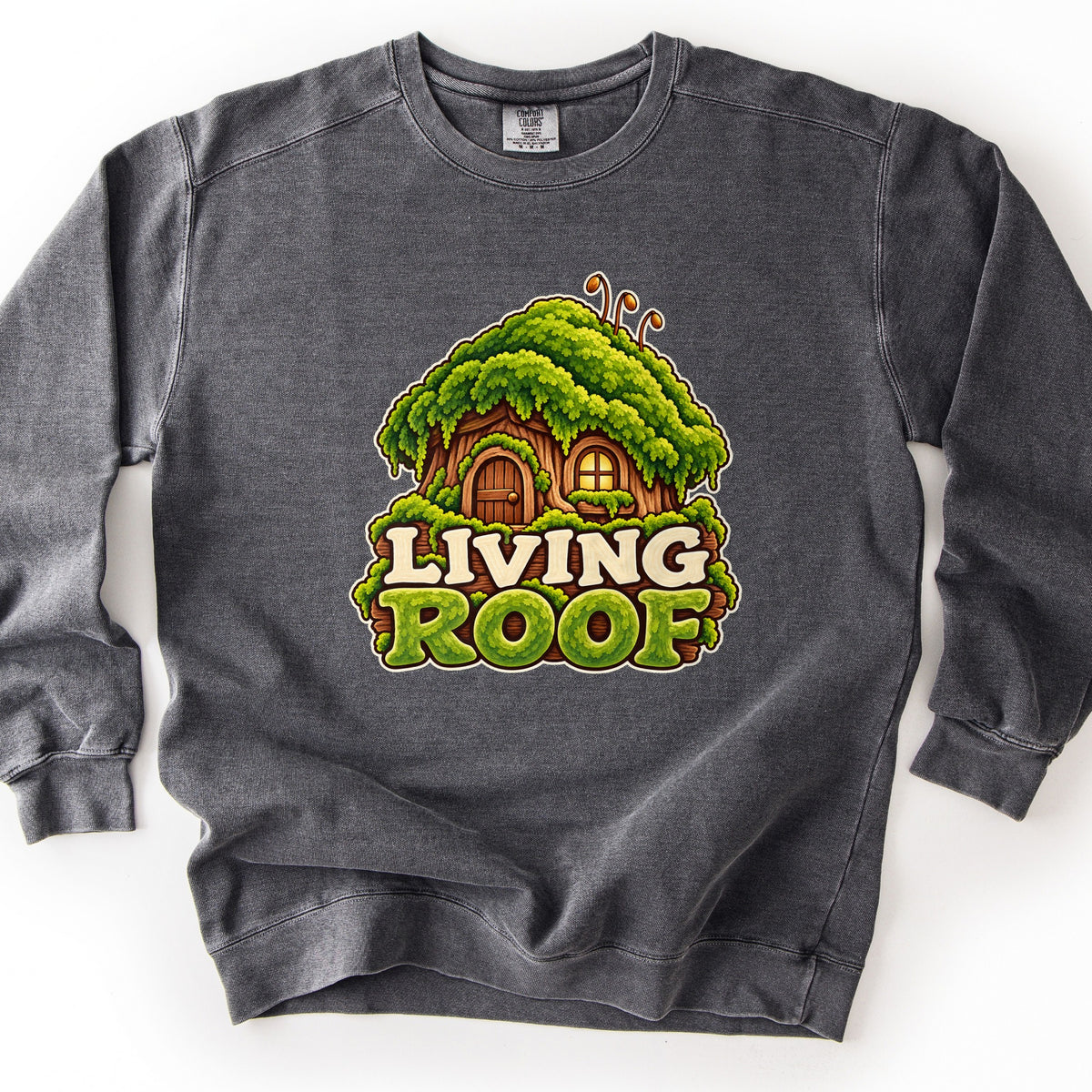 Living Roof Comfort Colors crewneck sweatshirt in Pepper, laid flat. A cartoon tree-stump cottage with a wooden door and glowing window under a thick moss roof sits above the words Living Roof, with Roof in mossy green letters. Shown on a white background.