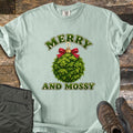 Merry and Mossy Comfort Colors T-shirt in Bay, laid flat. A round Christmas ornament made of green moss, topped with a gold cap and red bow, sits between the words Merry and Mossy in green lettering. Olive hiking pants and brown hiking boots lie beside it on weathered wood.