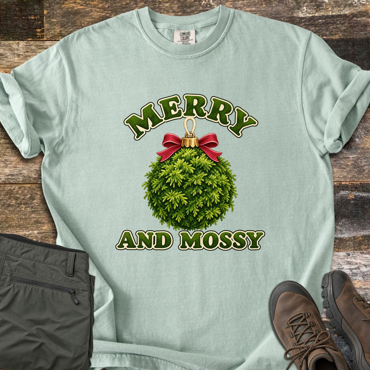 Merry and Mossy Comfort Colors T-shirt in Bay, laid flat. A round Christmas ornament made of green moss, topped with a gold cap and red bow, sits between the words Merry and Mossy in green lettering. Olive hiking pants and brown hiking boots lie beside it on weathered wood.